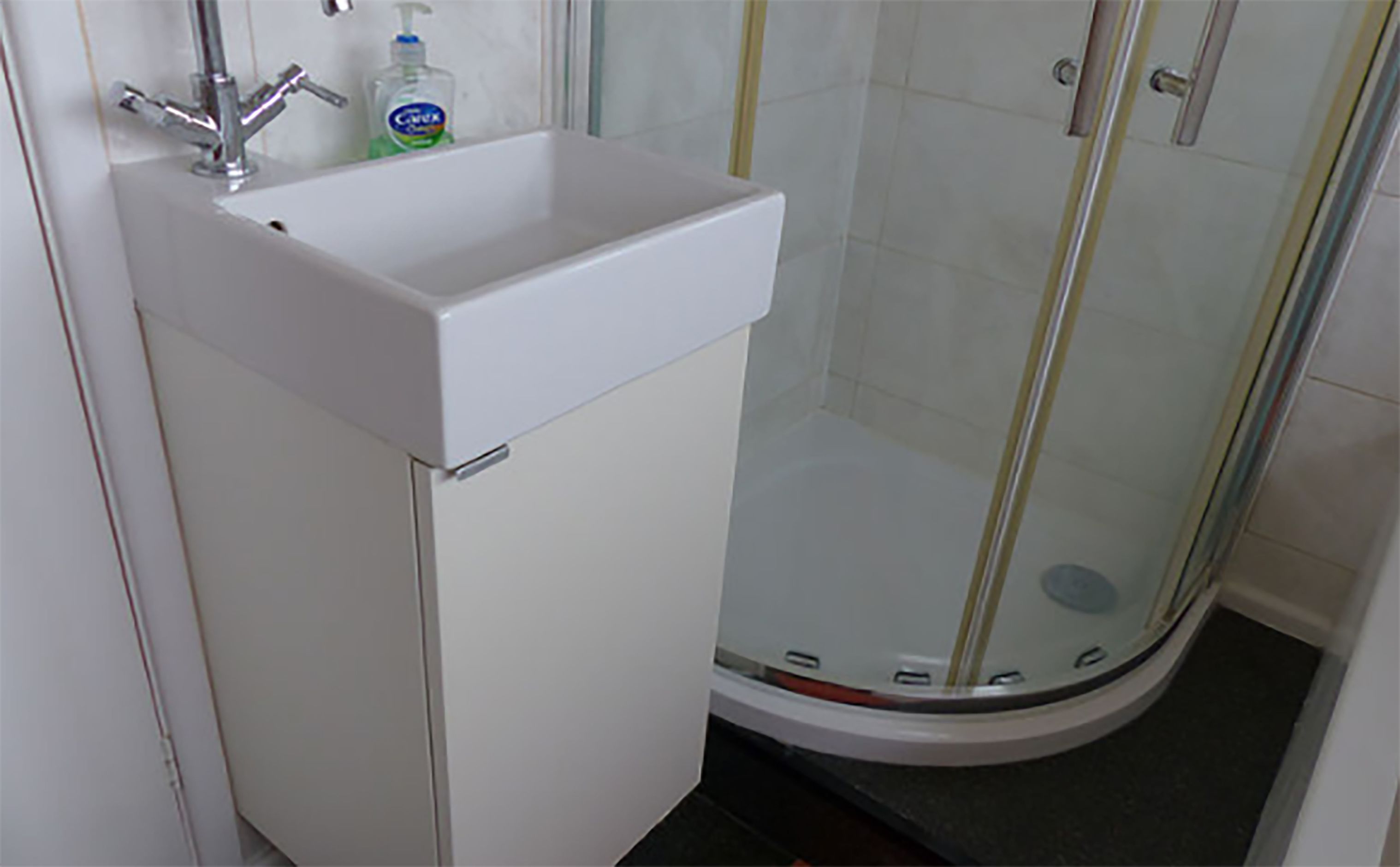 Un baño con lavamanos blanco, cabina de ducha con paredes de vidrio y una botella de jabón para manos.