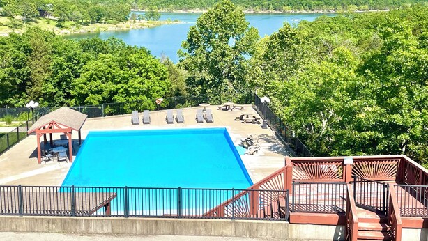 Ein Pool mit Liegestühlen und einer Laube, umgeben von üppiger Vegetation und Blick auf einen See und Hügel.