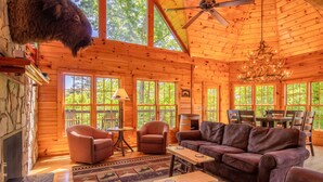 A cozy log cabin interior with a fireplace, leather furniture, and large windows offering a view of the outdoors. - Luxury Cabin, Casino, Ski, Snow Tube, Golf, Hike, Fish, Hot Tub, Fire Pit, Train (Whittier)