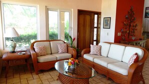 Ein Wohnzimmer mit zwei Sofas, einem Glastisch und einer Lampe. Eine Tür führt nach draußen, und ein kleiner Tisch trägt eine Lampe und eine Blumenvase.