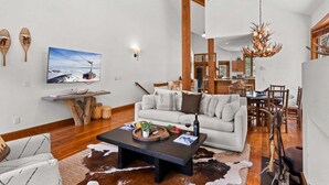 A spacious living room with a large sofa, a coffee table, and a television. There is a dining area with wooden chairs and a chandelier. - RMR: Tram Tower 4 Condo in Teton Village (Teton Village)