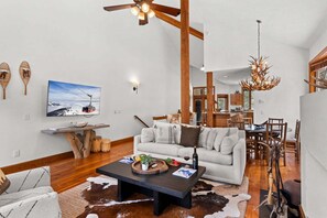 A spacious living room with a large sofa, a coffee table, and a television. There is a dining area with wooden chairs and a chandelier.