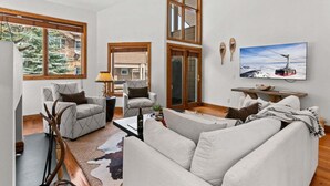 A living room with a sofa, armchairs, a coffee table, a TV, and a large window with a view of trees. - RMR: Tram Tower 4 Condo in Teton Village (Teton Village)