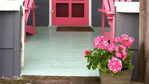 Un porche avec une porte rouge, deux fauteuils Adirondack et un pot de fleurs contenant des fleurs roses.