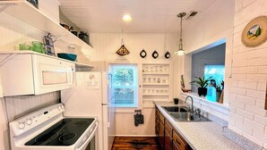 Une cuisine de taille réduite, dotée d’appareils électroménagers blancs, d’un plancher en bois et d’une fenêtre avec des stores.