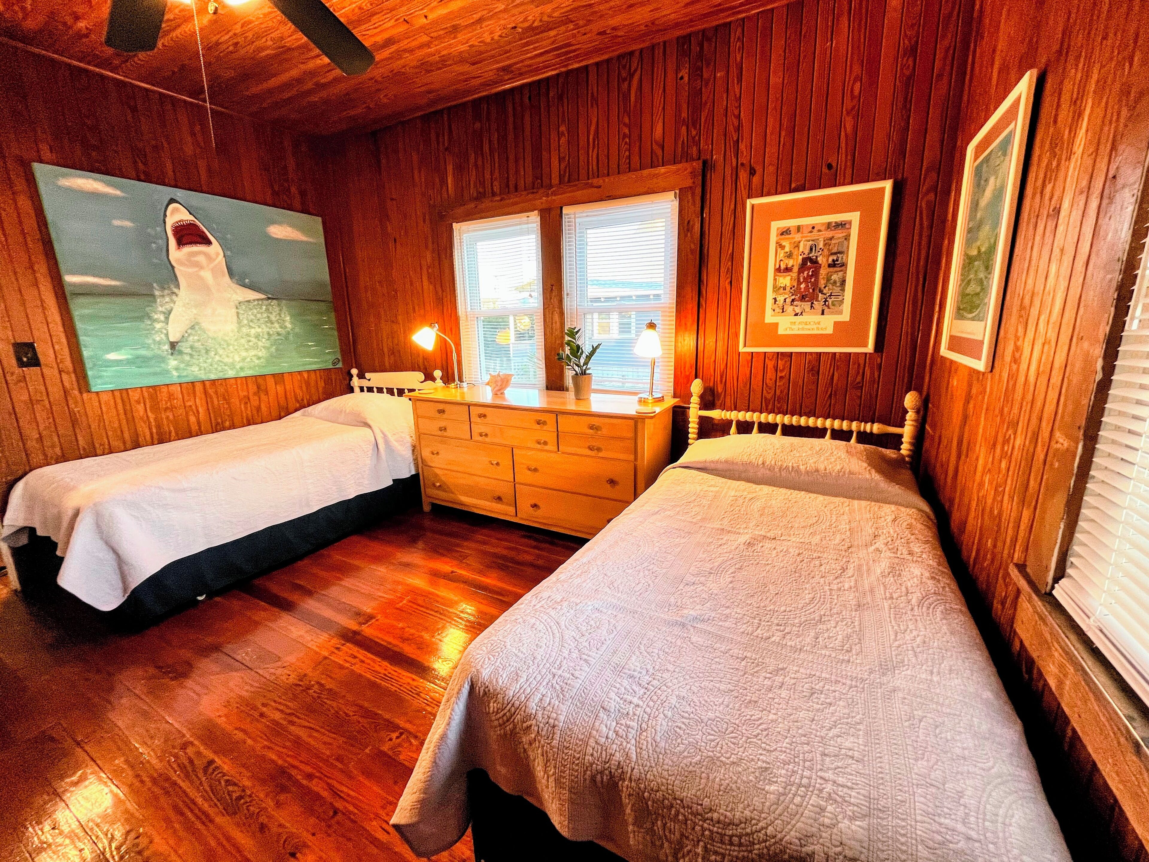 Een kamer met houten wanden en vloeren, twee bedden, een kapstok en een schilderij van een haai, plus een raam met zonwering.