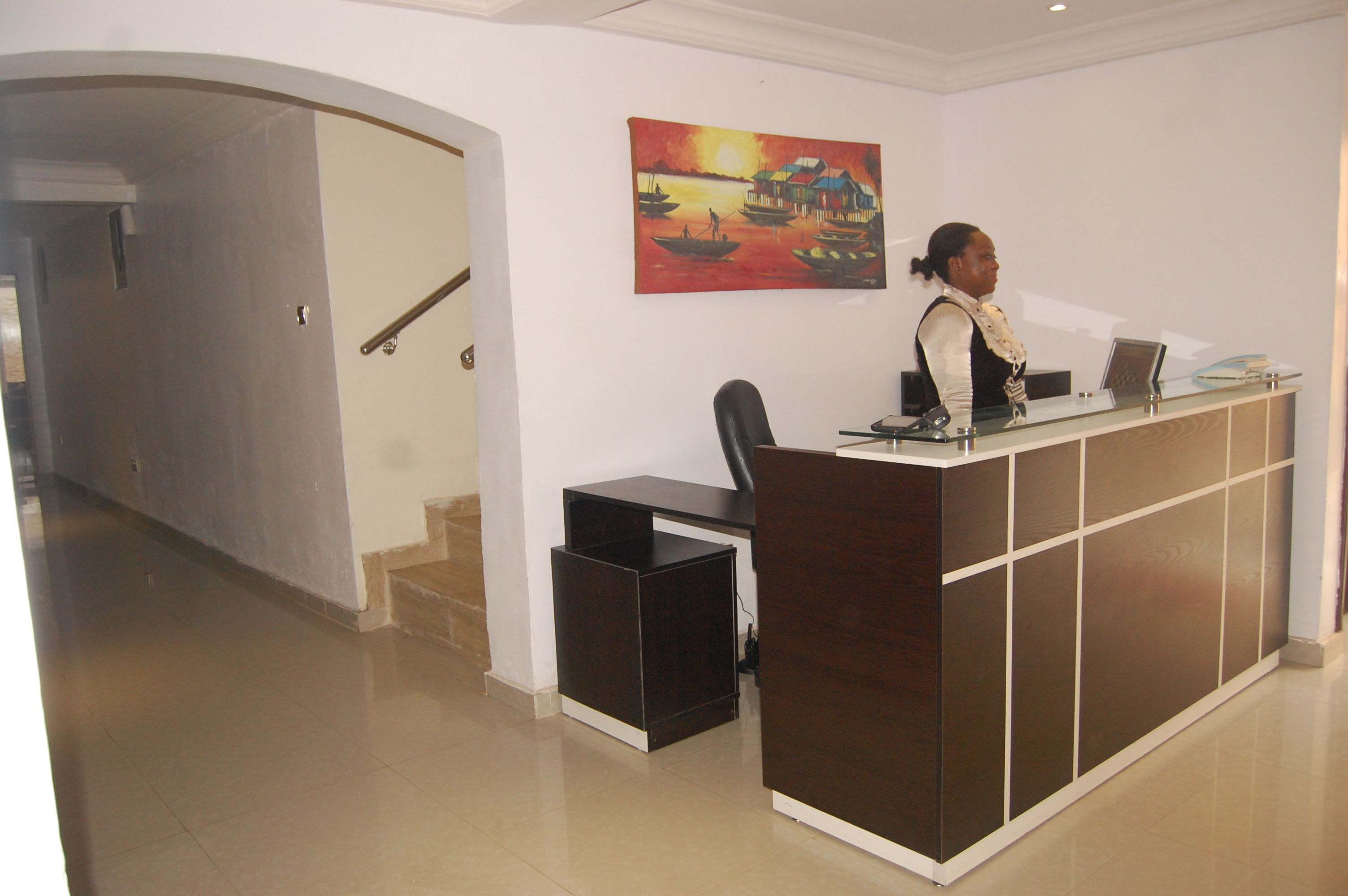 A reception desk with a person standing behind it, a painting on the wall, and a staircase leading to another area.