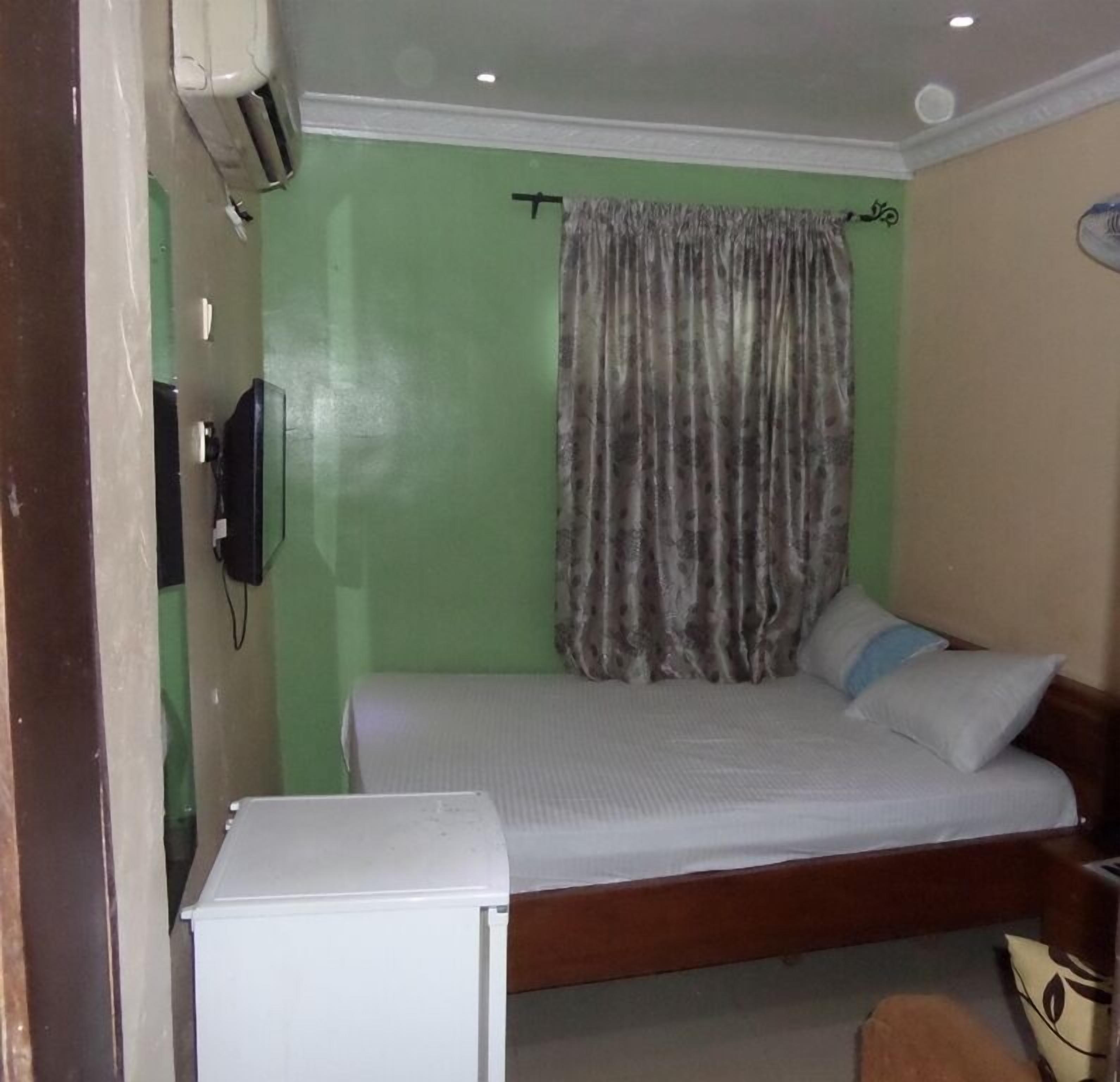 A small, single-bed room with a green wall, a wooden bed frame, a white bedside table, and a curtain.