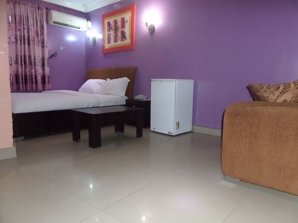 A room with a bed, a small table, a sofa, a refrigerator, and a wall-mounted light fixture.