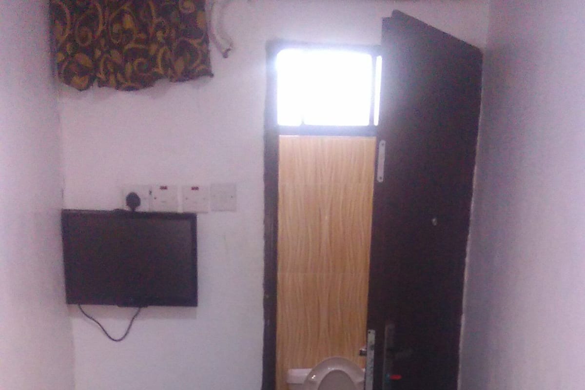 Quarto pequeno e estreito, com uma cama, mesa de cabeceira, televisão na parede e um toalete visível por uma porta aberta.