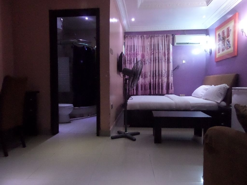 A bedroom with a bed, a standing fan, a window with curtains, and a wall-mounted air conditioner.