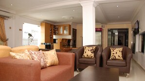 A living room with a brown sofa, a coffee table, and two armchairs with patterned cushions. There is a kitchen area with wooden cabinets and a dining table. - BEN AUTO guest house (Lagos)