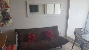 A living area with a brown sofa, two red cushions, a white-framed mirror, a television, and a glass table with chairs. - Departamento Euromarina (Vina del Mar)