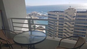 A balcony with a round glass table and wicker chairs, overlooking a rocky coastline and a building with multiple floors. - Departamento Euromarina (Vina del Mar)