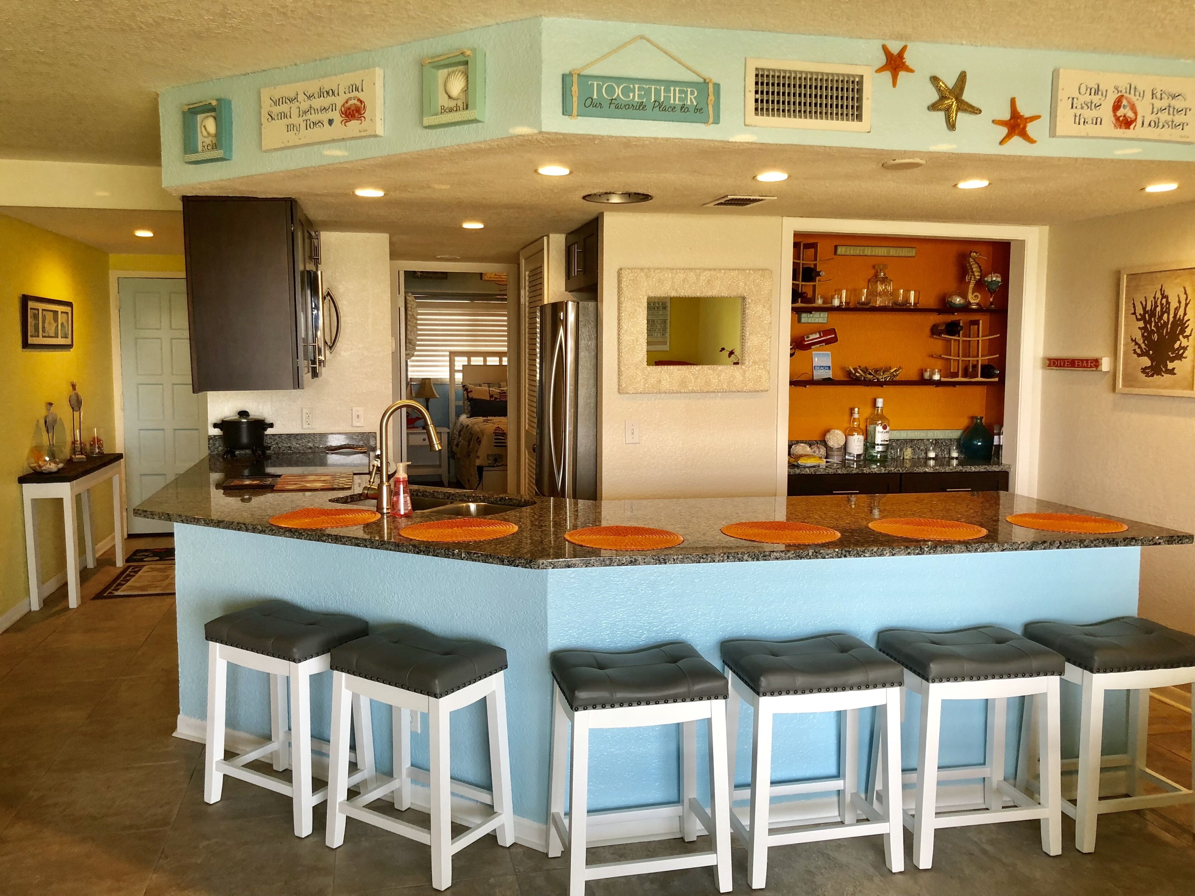 Una cocina con una isla central, taburetes de bar blancos y decoración mural.