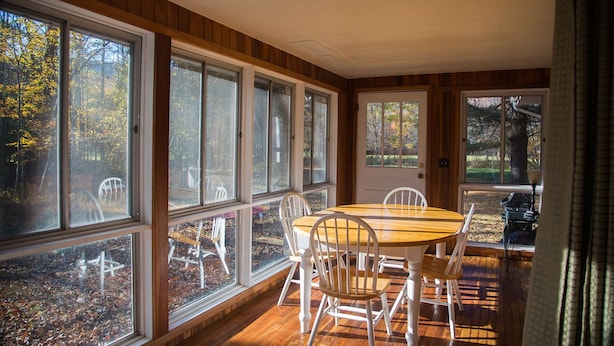 A dining area with a wooden table and white chairs, large windows with a view of trees, and a door leading outside.