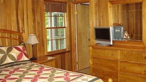 A bedroom with a wooden headboard, a bed with a quilt, a bedside lamp, a dresser, a television, and a window with blinds.