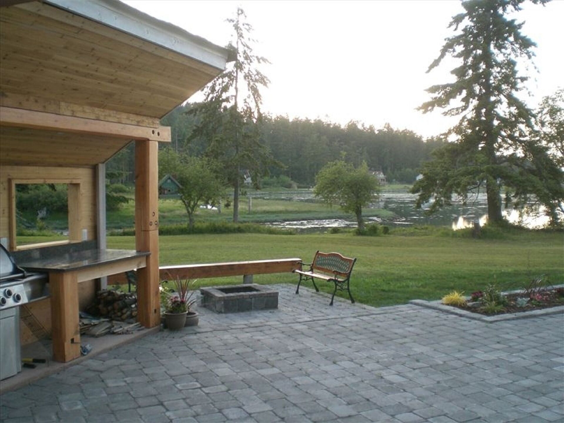 A patio area with a wooden deck, a grill, and a bench overlooking a river and forested area.