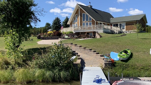 Una casa con un muelle que se adentra en un lago, rodeada de vegetación y un cielo despejado.