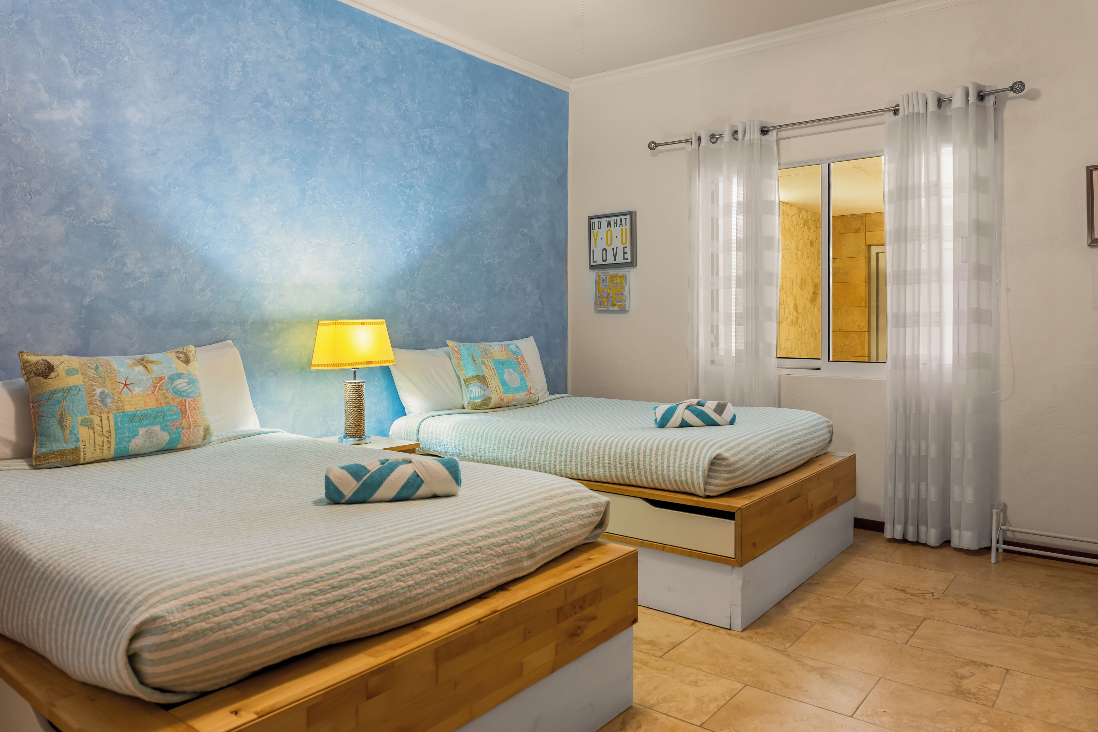 Ein Schlafzimmer mit zwei Betten, einem Fenster mit Vorhängen, einer Wanduhr und einem kleinen Tisch mit einer Lampe.