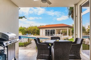 Une terrasse avec un barbecue, des chaises en osier, une table et une vue sur une piscine et une maison.