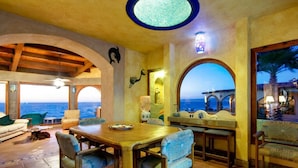 Zona pranzo con vista sul mare, caratterizzata da un tavolo e sedie in legno, una ventola a soffitto e un lampadario.
