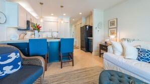 A modern kitchen with white cabinets, a black refrigerator, and blue bar stools. There is a dining area with a blue sofa and a coffee table. The living room features a white sofa with blue and white patterned pillows. - Gorgeous remodeled condo Oceanfront! SUNSET! <br> (Kailua-Kona)