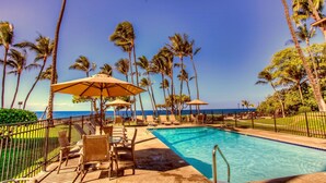 A poolside area with lounge chairs and umbrellas, surrounded by palm trees and a view of the ocean. - Gorgeous remodeled condo Oceanfront! SUNSET! <br> (Kailua-Kona)