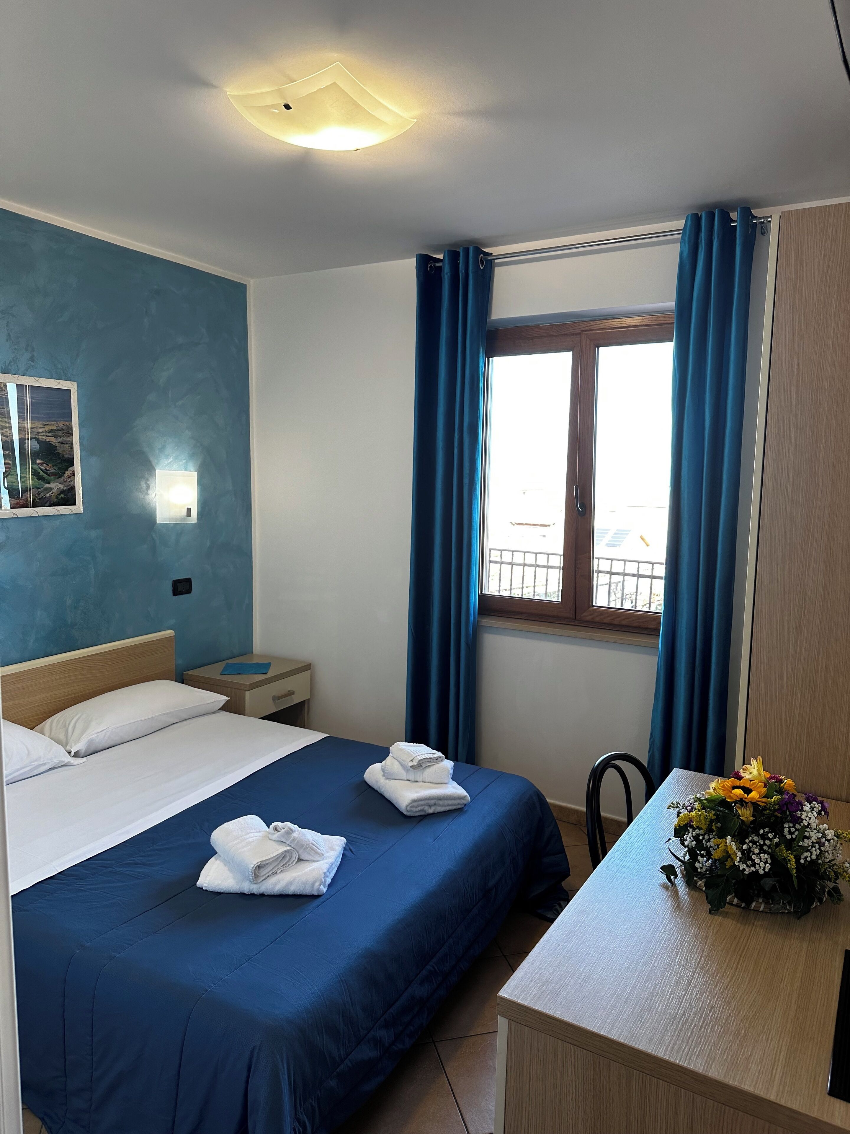 Habitación de hotel con cama, escritorio, ventana con cortinas, mesita de noche y lámpara en la pared.