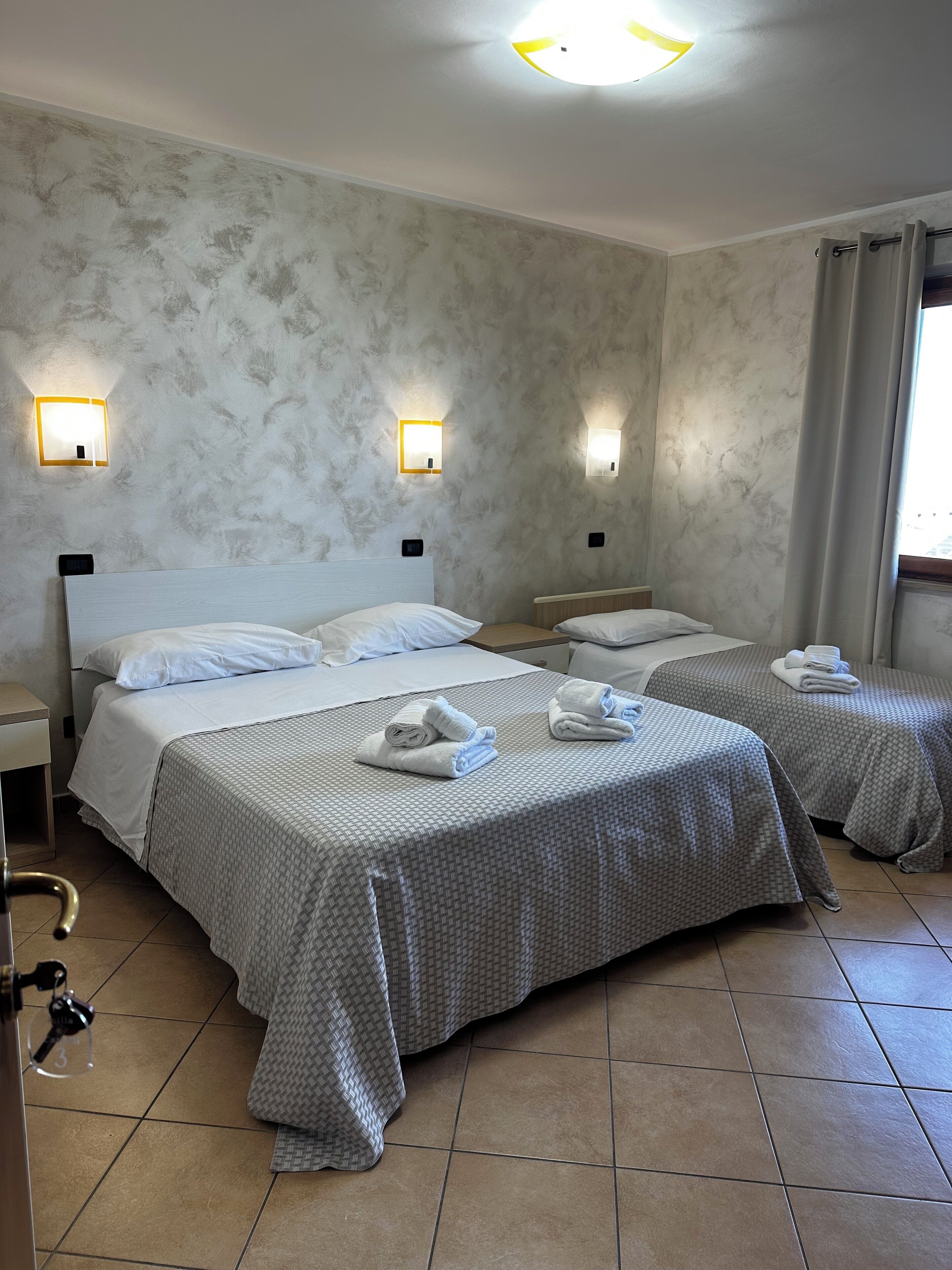 Quarto de hotel com duas camas, cada uma com cabeceira branca e edredons cinza estampados. Há luminárias fixadas na parede e uma luminária no teto.