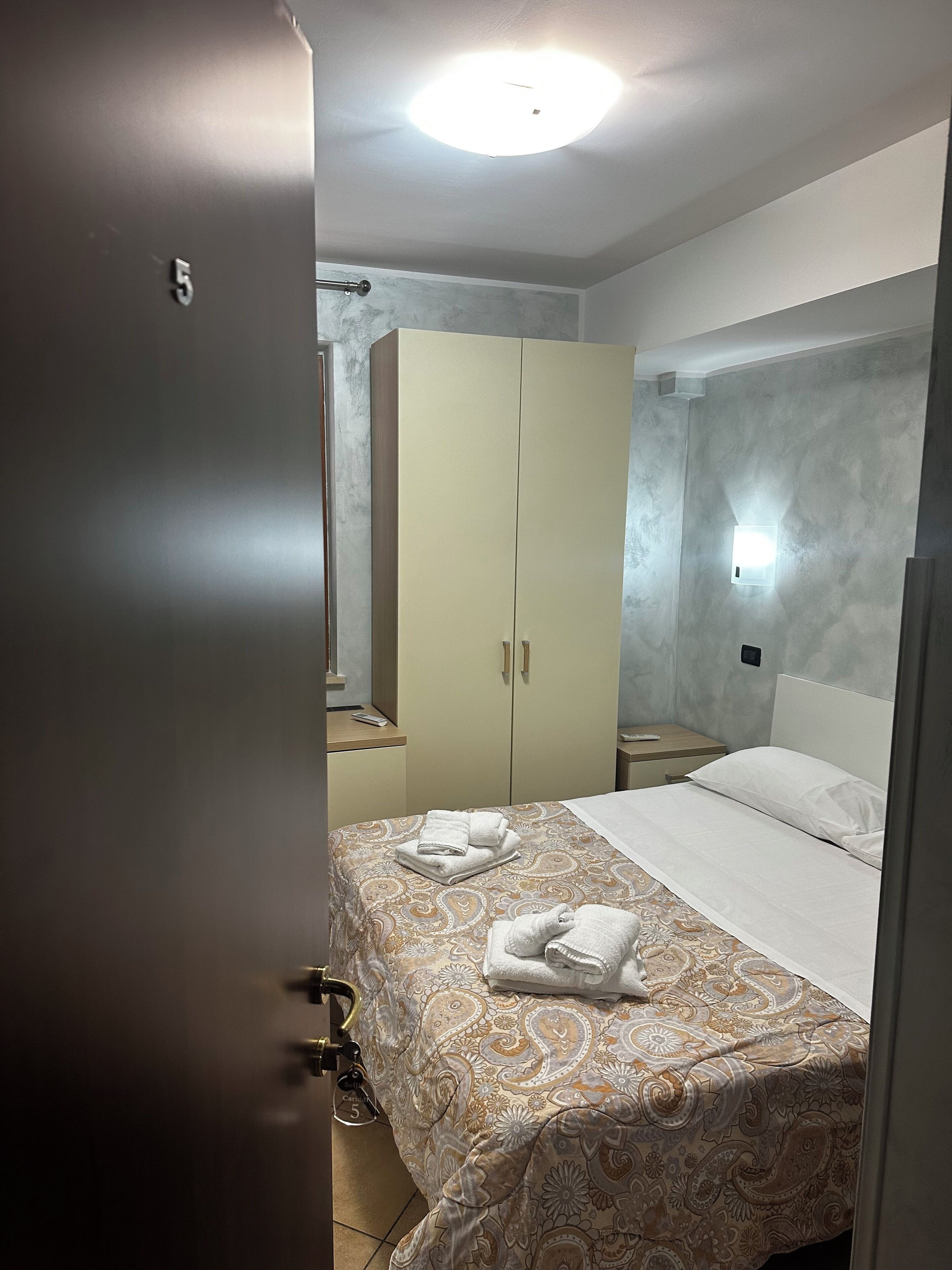 A hotel room with a bed, a wardrobe, a nightstand, and a wall-mounted light.