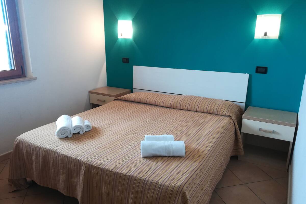 A neatly made bed with striped bedding and two rolled towels, a bedside table with a drawer, and a wall-mounted light fixture.