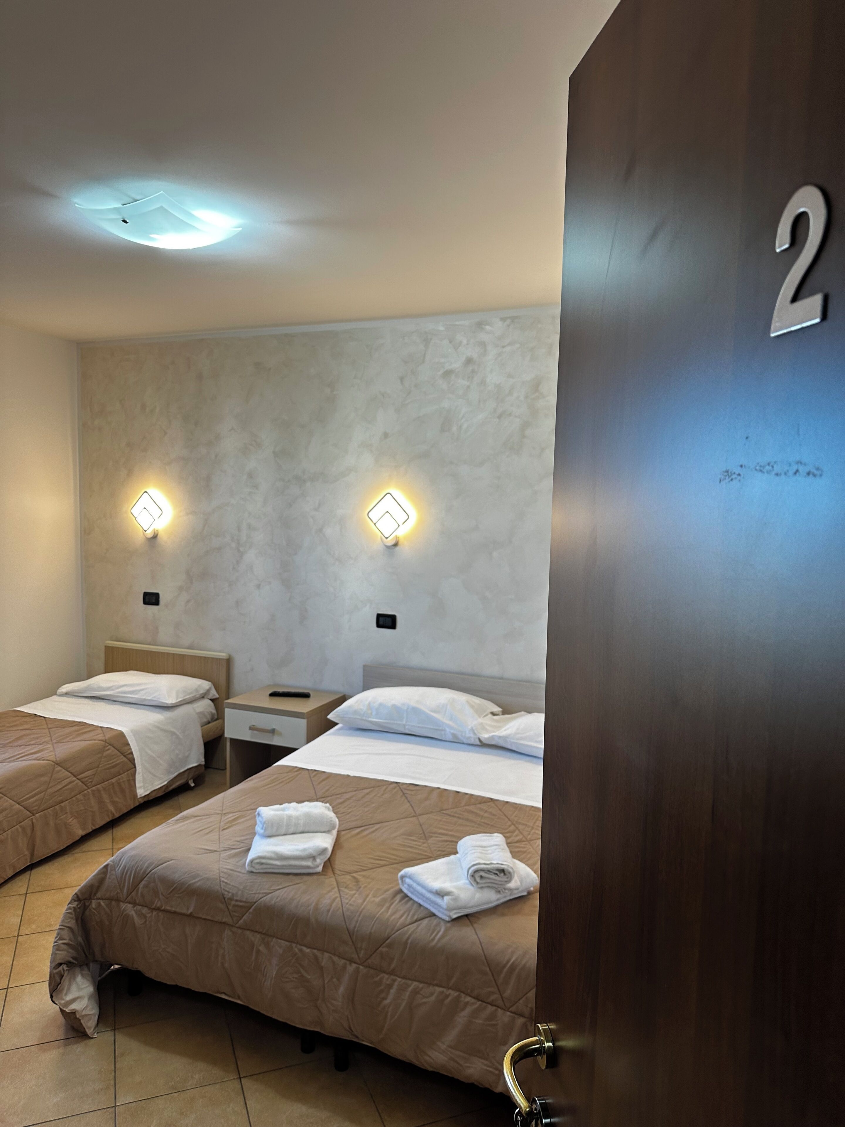 A hotel room with two beds, each with a headboard, a nightstand, and a wall-mounted light. The room has a textured wall and a door with the number 2.