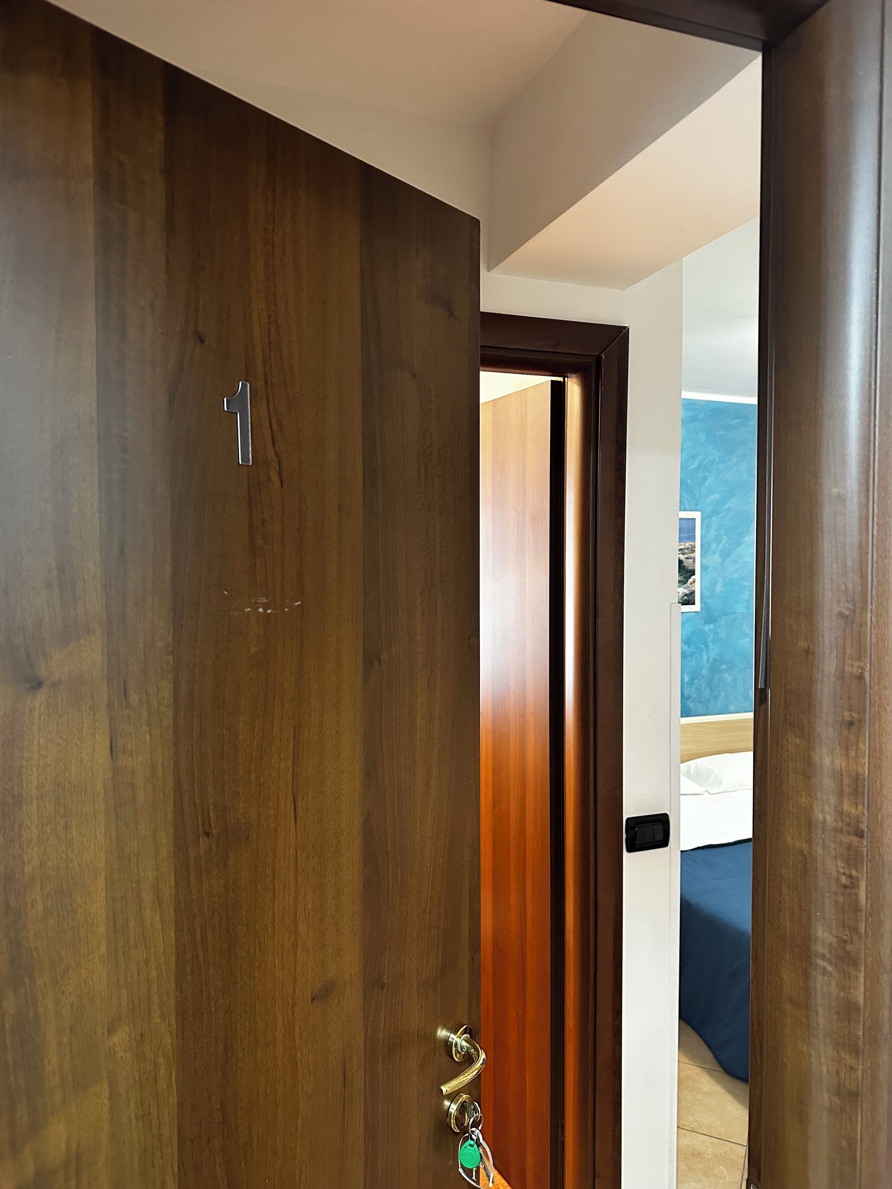 Una puerta de madera con un ojo de cerradura y cerradura, que da a una habitación con una cama y una pared con un diseño azul texturizado.
