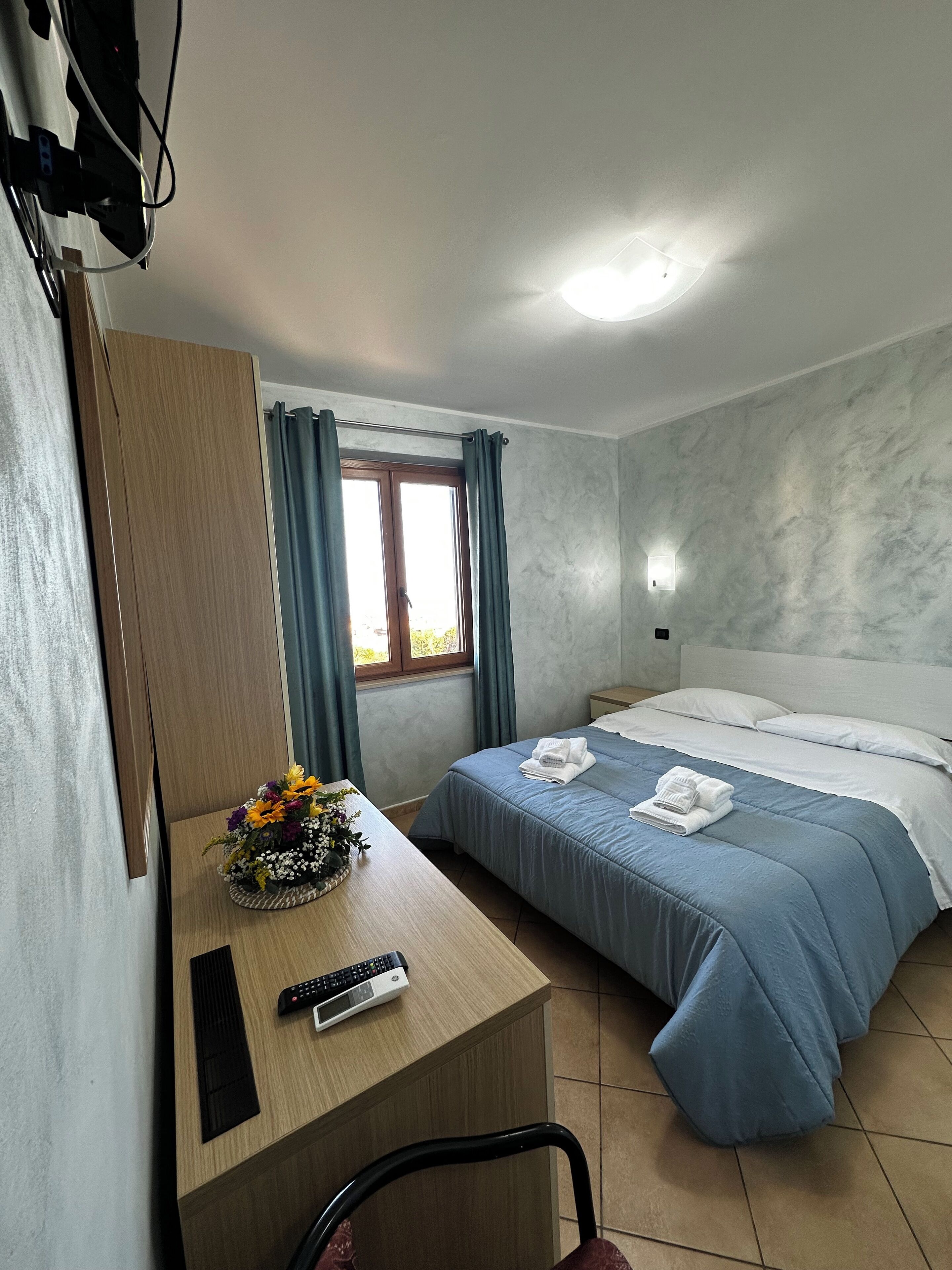 Ein Hotelzimmer mit Bett, Schreibtisch mit Fernbedienung, Stuhl, Fenster mit Vorhängen und einer Wandleuchte.