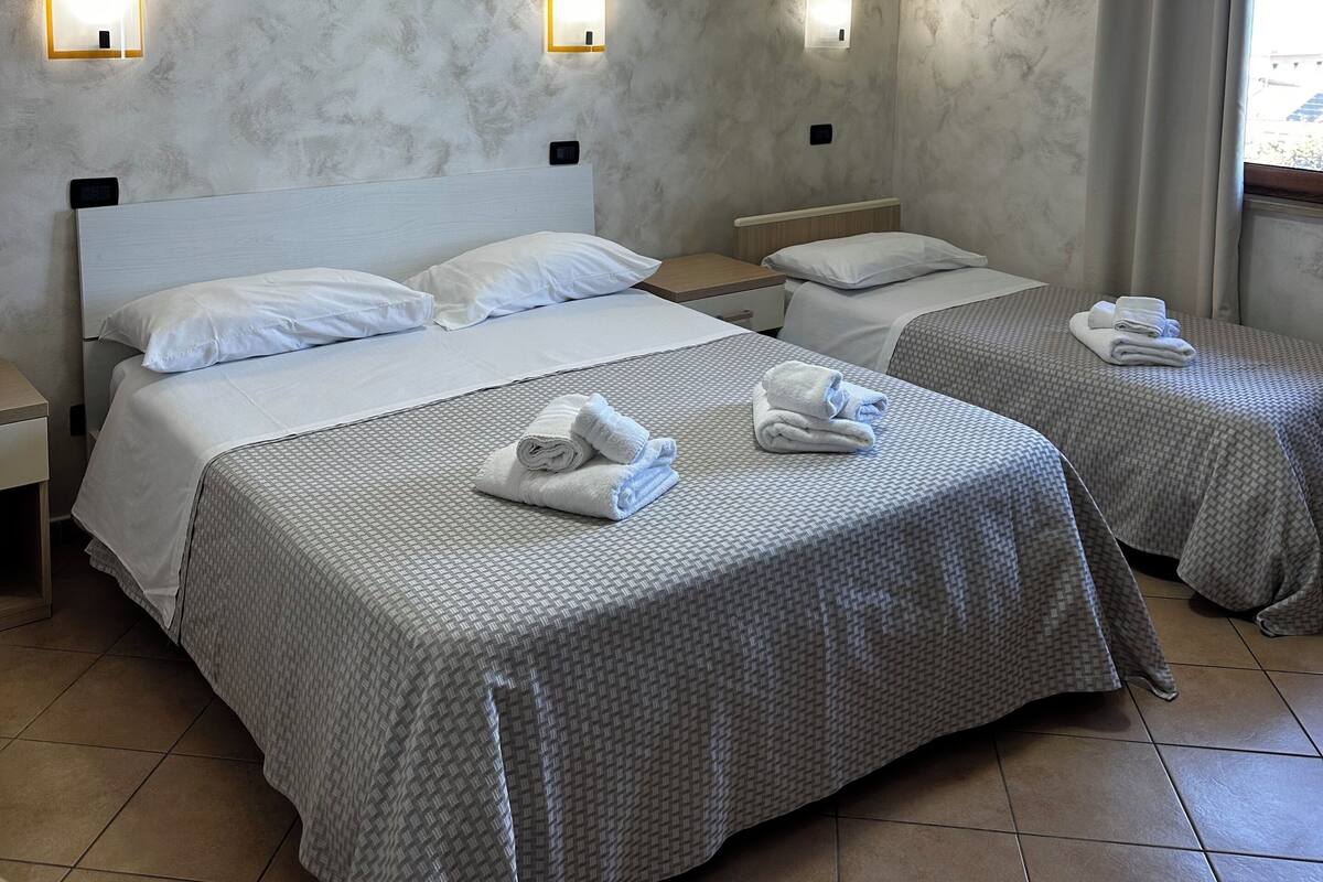 A hotel room with two beds, each with a patterned bedspread, white pillows, and neatly folded towels on the beds. There are wall-mounted light fixtures and a ceiling light.
