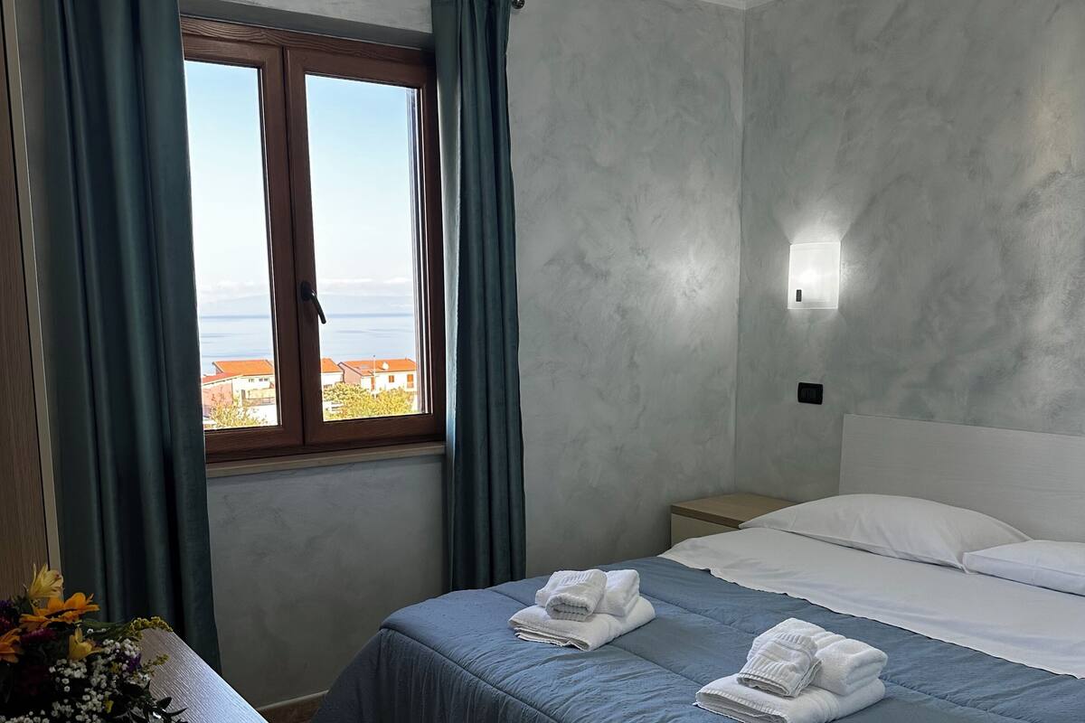 A hotel room with a bed, a desk with a remote control, a window with curtains, and a wall-mounted light.