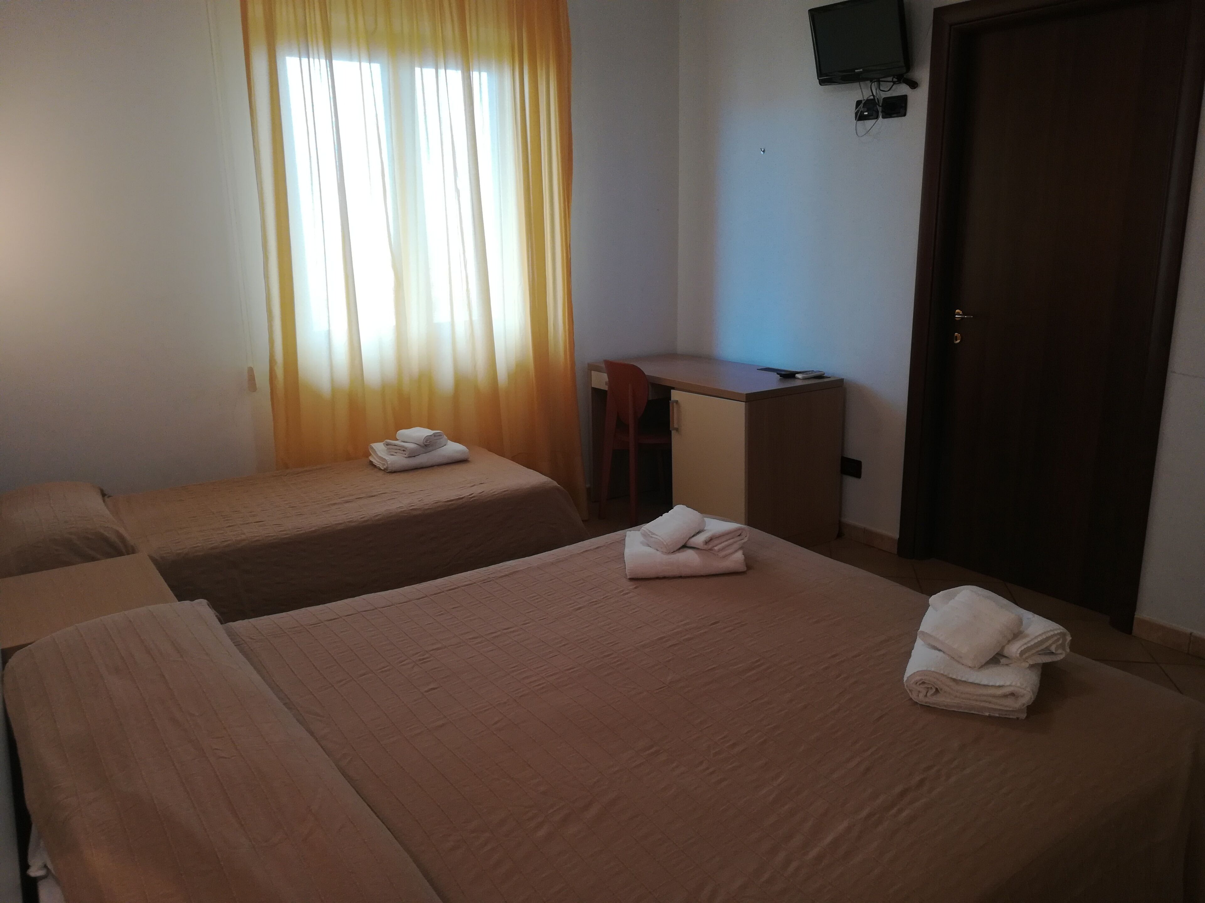 Ein Hotelzimmer mit zwei Einzelbetten, einem Schreibtisch, einem Stuhl und einem an der Wand befestigten Fernseher.