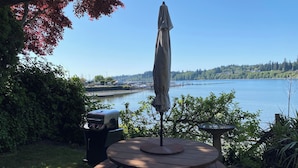 Taula de picnic de fusta amb una ombrel·la, un barbacoa i vista d'un llac amb barques i un moll al fons.