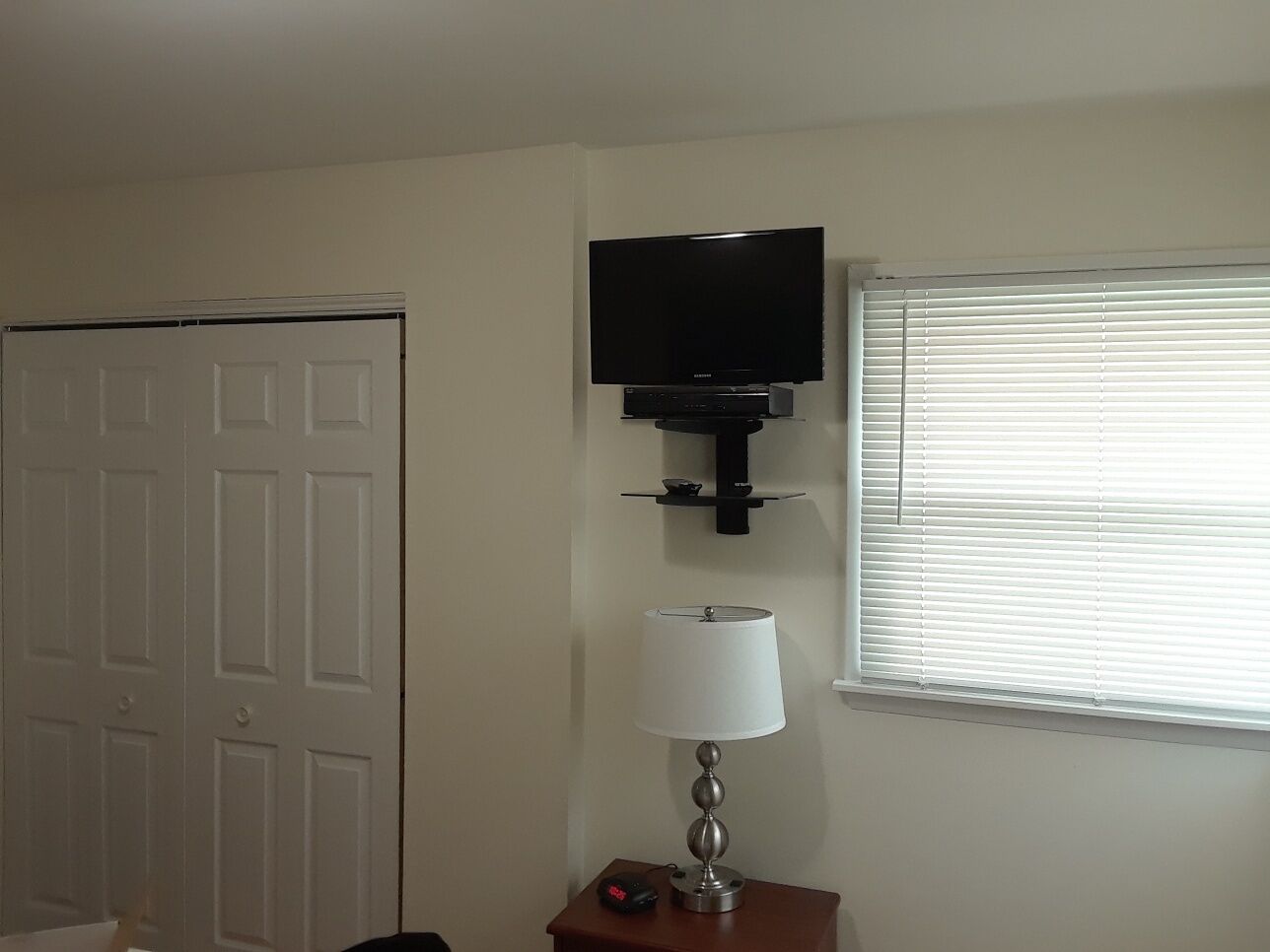 A room with a closed door, a flat-screen TV mounted on the wall, a lamp on a small table, and a window with blinds.