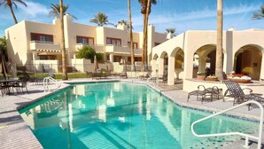 A swimming pool with clear blue water, surrounded by lounge chairs and umbrellas, with a building featuring arches and palm trees in the background.