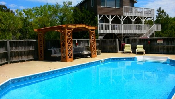 Une piscine agrémentée d’une pergola en bois et de chaises longues, une terrasse, et une maison avec un balcon en arrière-plan.