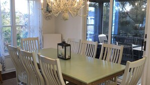 Une salle à manger avec une grande table entourée de chaises blanches, un lustre et de grandes fenêtres donnant sur l’extérieur.