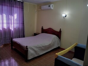 A bedroom with a bed, a nightstand, a chair, a window with curtains, and an air conditioning unit. - Appartement (Grand-Baie)