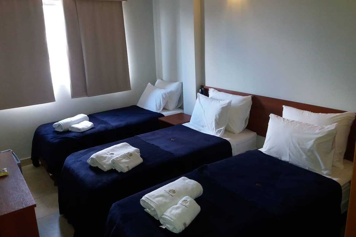 A hotel room with two beds, each with a blue bedspread and white pillows, and a wooden headboard. There is a nightstand with a lamp and a small table with a bottle and a glass.