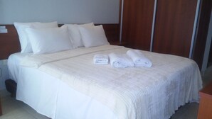 Cama bem arrumada com lençóis e almofadas brancos, uma toalha dobrada e cabeceira de madeira.