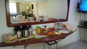 Uma mesa de café da manhã com máquinas de café, acompanhamentos, frutas e um espelho refletindo uma área de refeições.