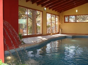 Piscina interna com grandes janelas, vigas de madeira e parede de pedra.
