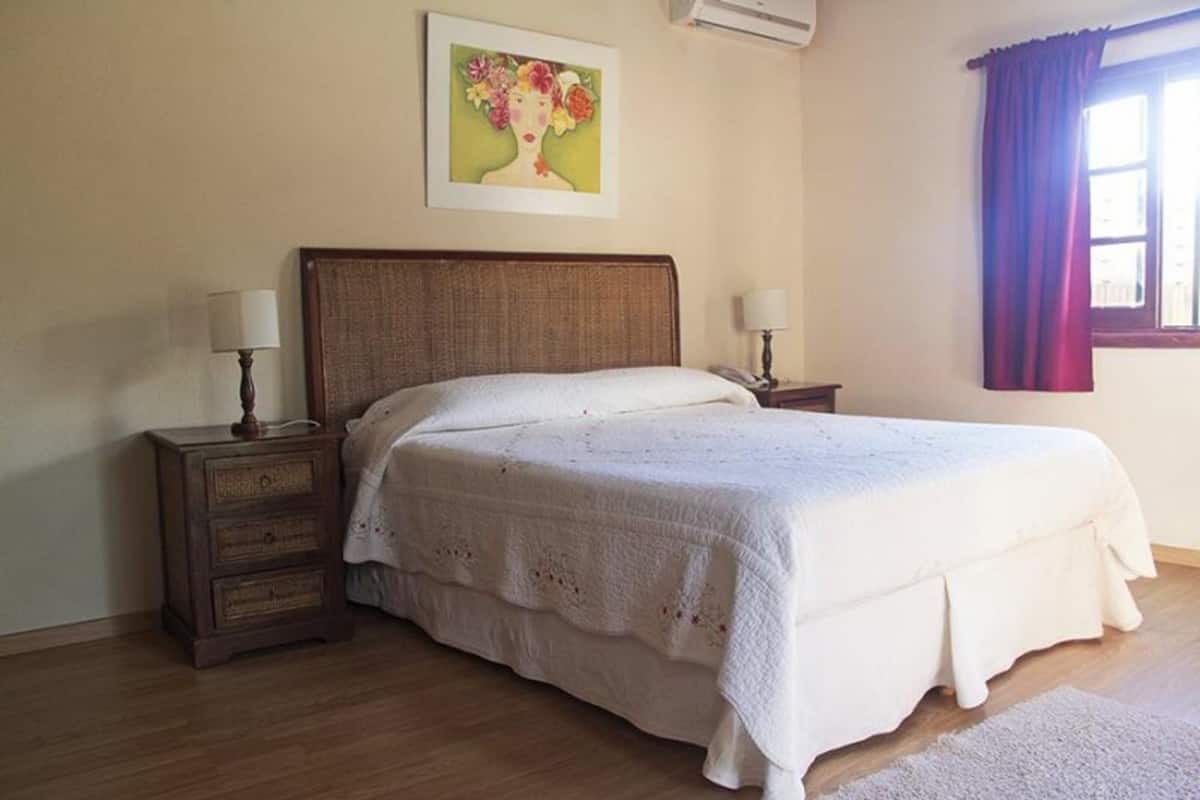 A bedroom with a wooden bed, two bedside tables with lamps, a painting on the wall, and a window with curtains.