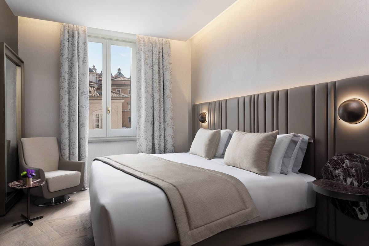 Et moderne hotelværelse med en stor seng, et siddeområde med en stol, et lille natbord og udsigt over byen gennem et vindue.